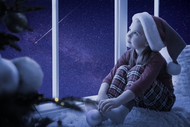 Cute little girl in Santa hat sitting on windowsill and looking at shooting star in beautiful night sky