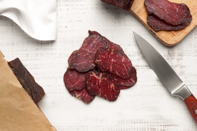 Photo of Pieces of delicious beef jerky and knife on white wooden table, flat lay