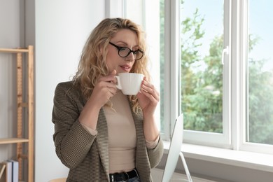 Beautiful young woman with cup of coffee near window in office