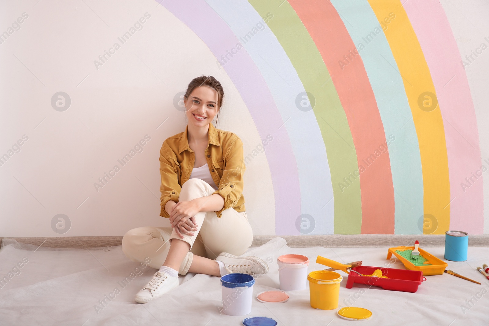 Photo of Young woman and decorator's tools near wall with painted rainbow indoors