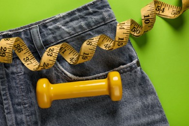 Photo of Jeans, dumbbell and measuring tape on green background, flat lay. Weight loss concept