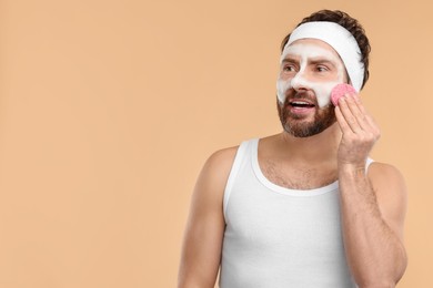 Photo of Man with headband washing his face using sponge on beige background, space for text