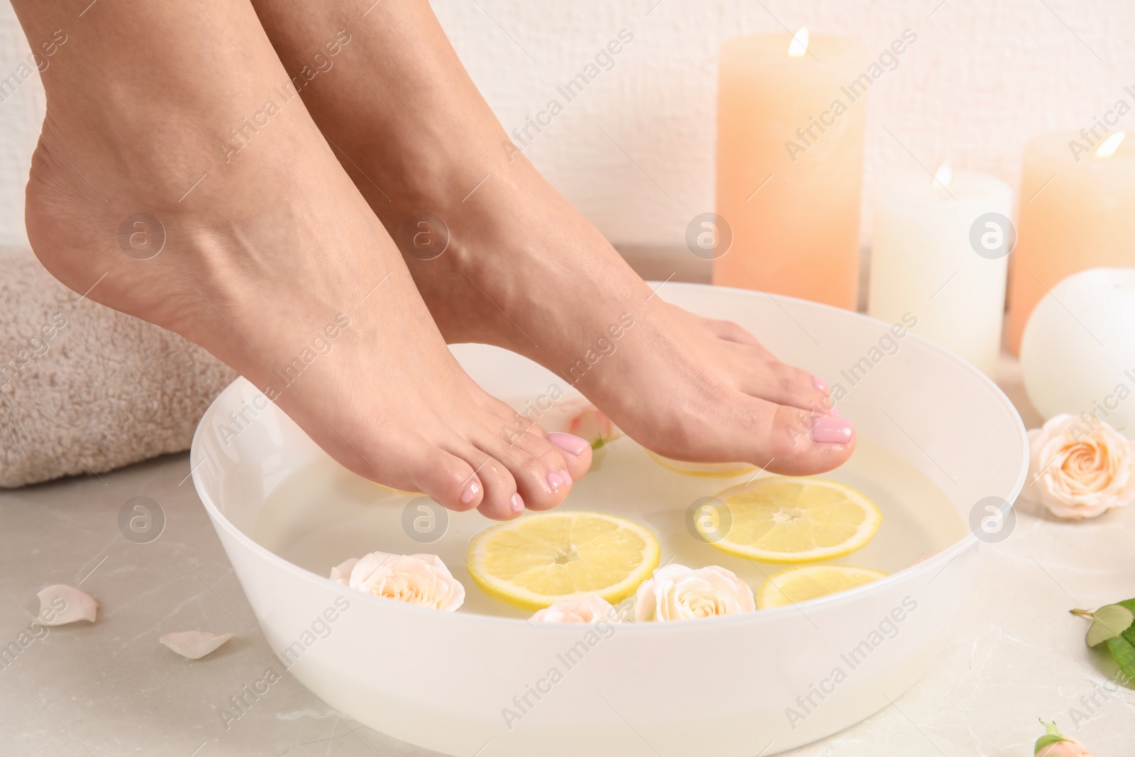 Photo of Woman putting her feet into bowl with water, roses and lemon slices on floor, closeup. Spa treatment