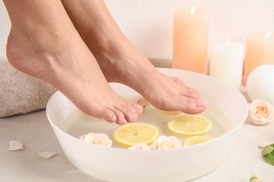 Photo of Woman putting her feet into bowl with water, roses and lemon slices on floor, closeup. Spa treatment