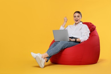 Happy woman with laptop sitting on beanbag chair against orange background. Space for text