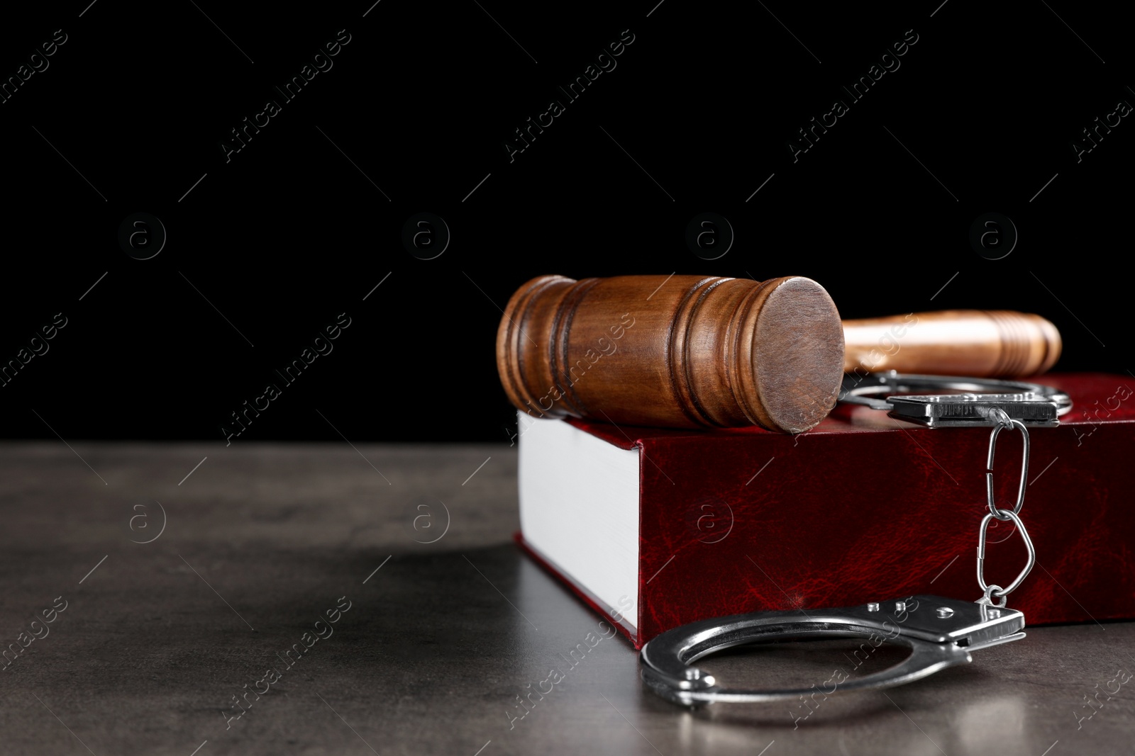 Photo of Judge's gavel, handcuffs and book on grey table against black background, space for text. Criminal law concept