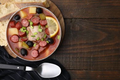 Photo of Meat solyanka soup with thin dry smoked sausages in bowl and bread slices on wooden table, flat lay