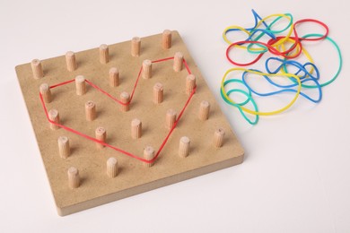 Photo of Wooden geoboard with heart made of rubber bands on white table. Educational toy for motor skills development
