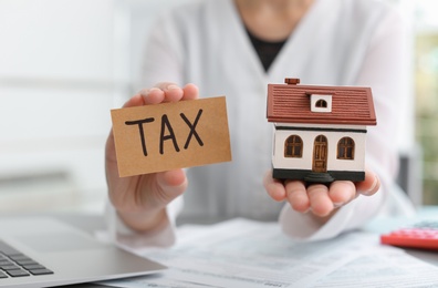 Photo of Woman holding house model and card with word TAX, closeup