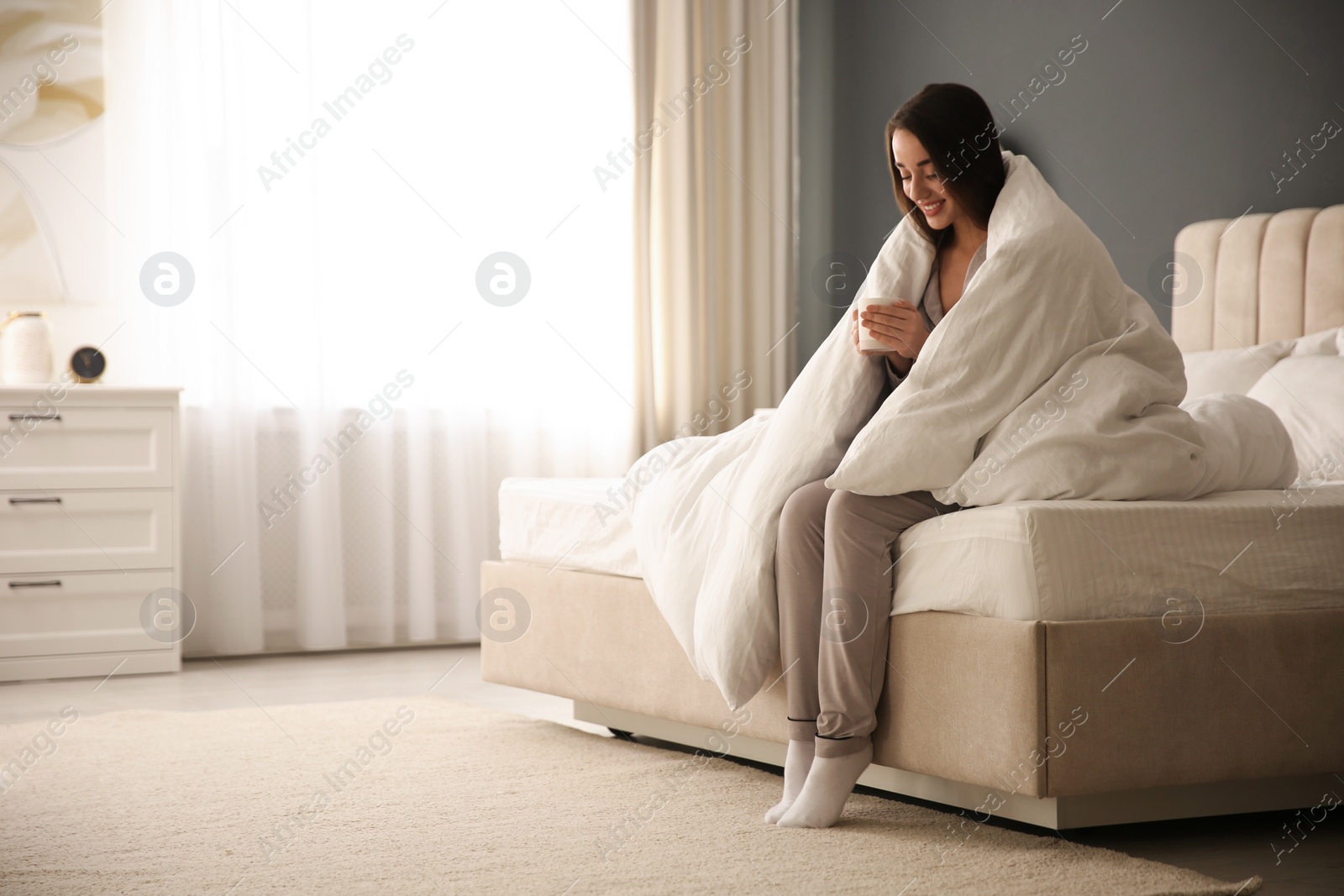 Photo of Beautiful young woman wrapped with soft blanket holding cup of hot drink on bed at home