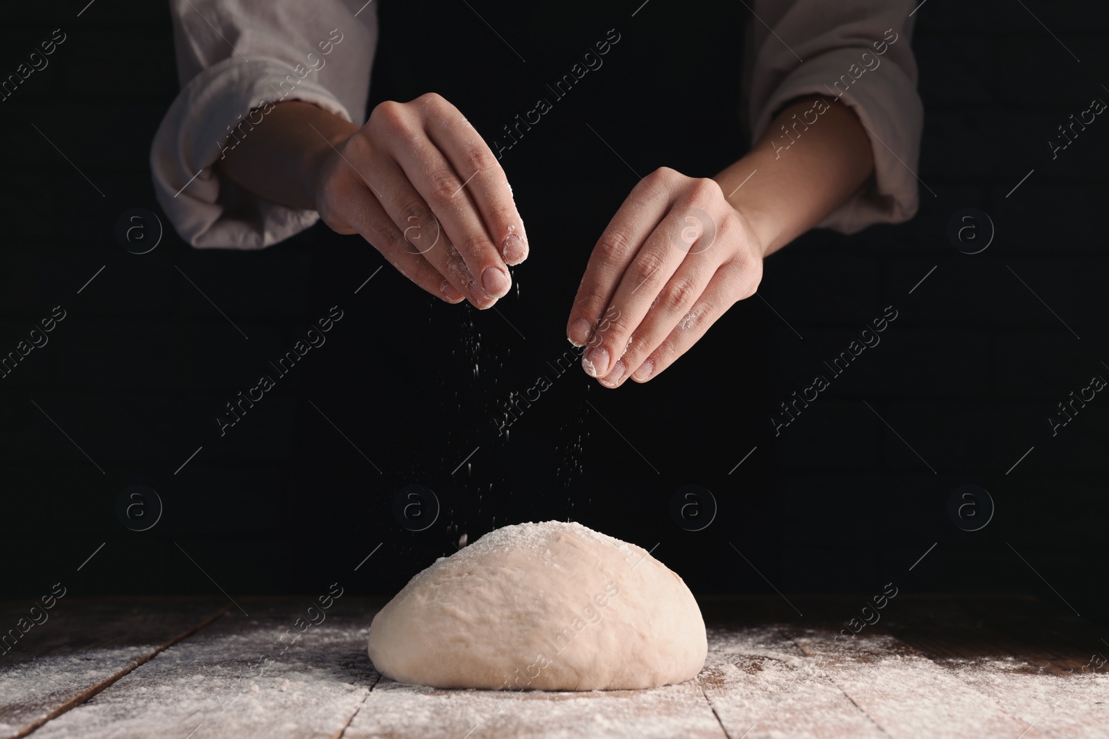 Photo of Woman sprinkling flour over dough at wooden table on dark background, closeup