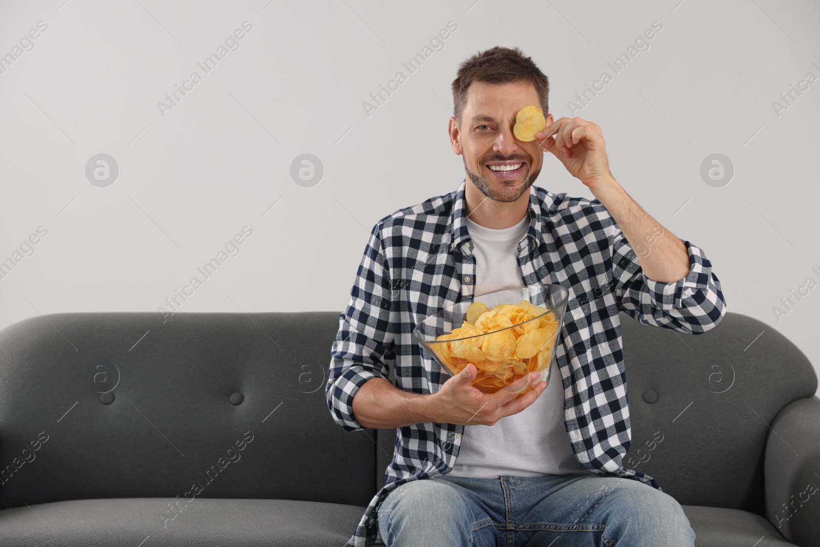 Photo of Handsome man holding potato chips near eye on sofa