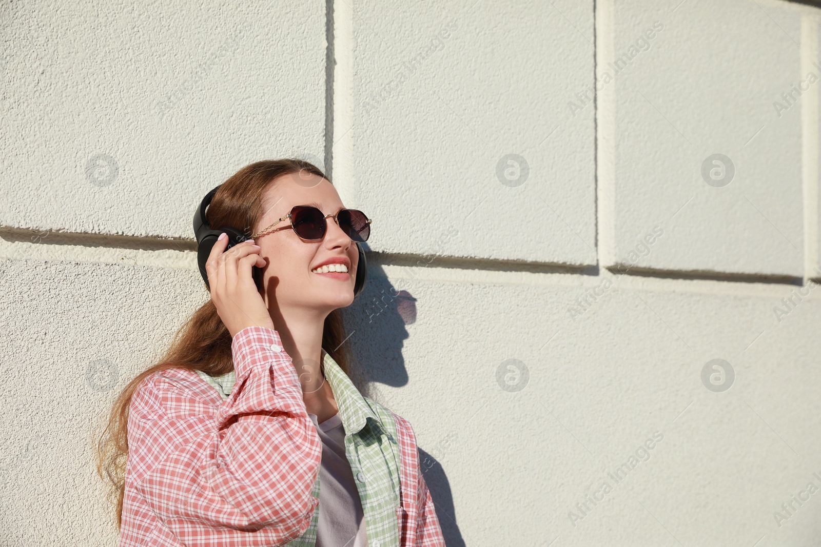 Photo of Smiling woman in headphones listening to music near white wall outdoors. Space for text