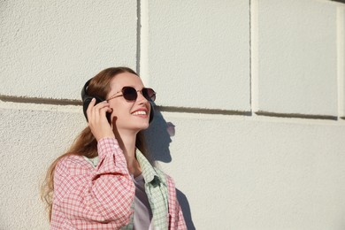 Smiling woman in headphones listening to music near white wall outdoors. Space for text