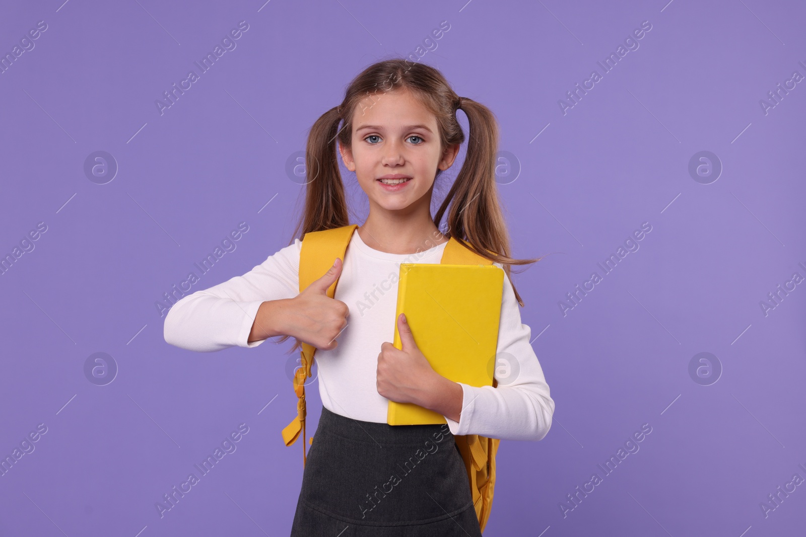 Photo of Smiling schoolgirl with backpack and book showing thumbs up on violet background