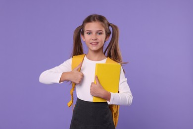 Smiling schoolgirl with backpack and book showing thumbs up on violet background