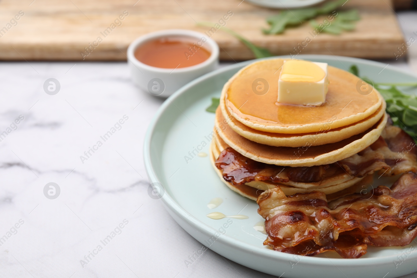 Photo of Tasty pancakes with butter, fried bacon and fresh arugula on white marble table, closeup. Space for text