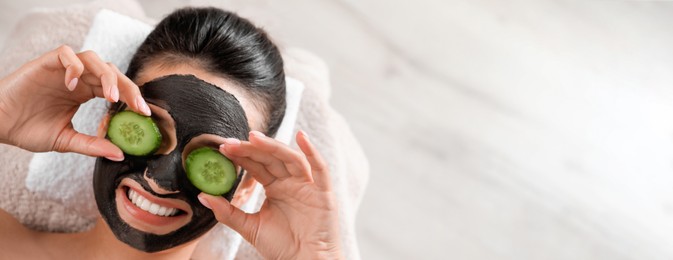 Beautiful woman with black mask on face and cucumber slices relaxing in spa salon, top view with space for text. Banner design