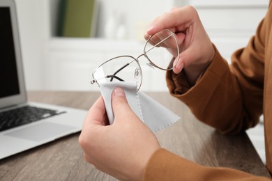Photo of Woman wiping her glasses with microfiber cloth at wooden table in office, closeup