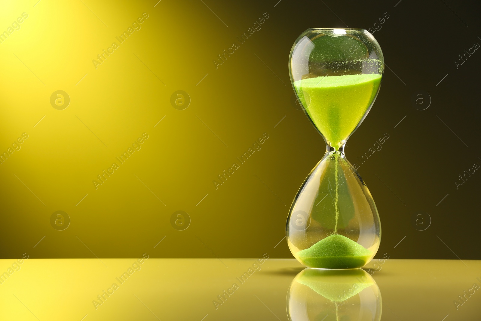 Photo of Hourglass with light green flowing sand on table against color background. Space for text