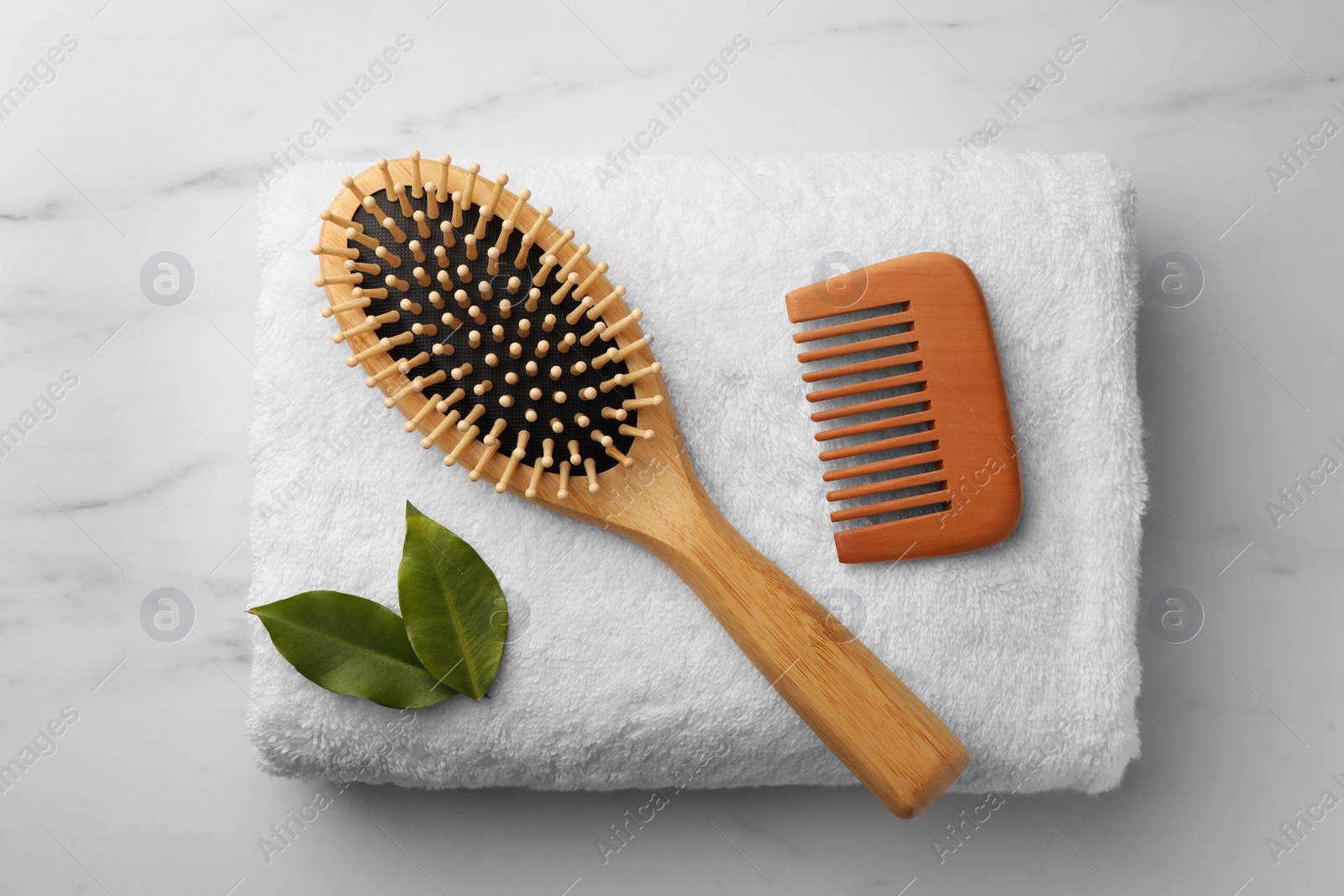 Photo of Wooden hair comb, brush, terry towel and green leaves on white marble table, top view