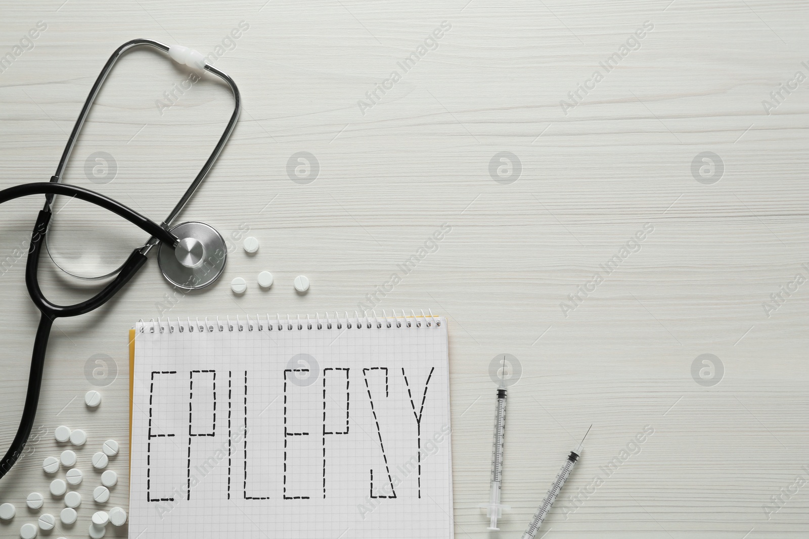 Photo of Notebook with word Epilepsy, stethoscope, pills and syringes on white wooden table, flat lay. Space for text