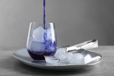 Pouring delicious blue matcha tea into glass with ice cubes on grey table, space for text