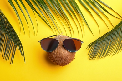 Coconut with sunglasses and palm leaves on yellow background, flat lay