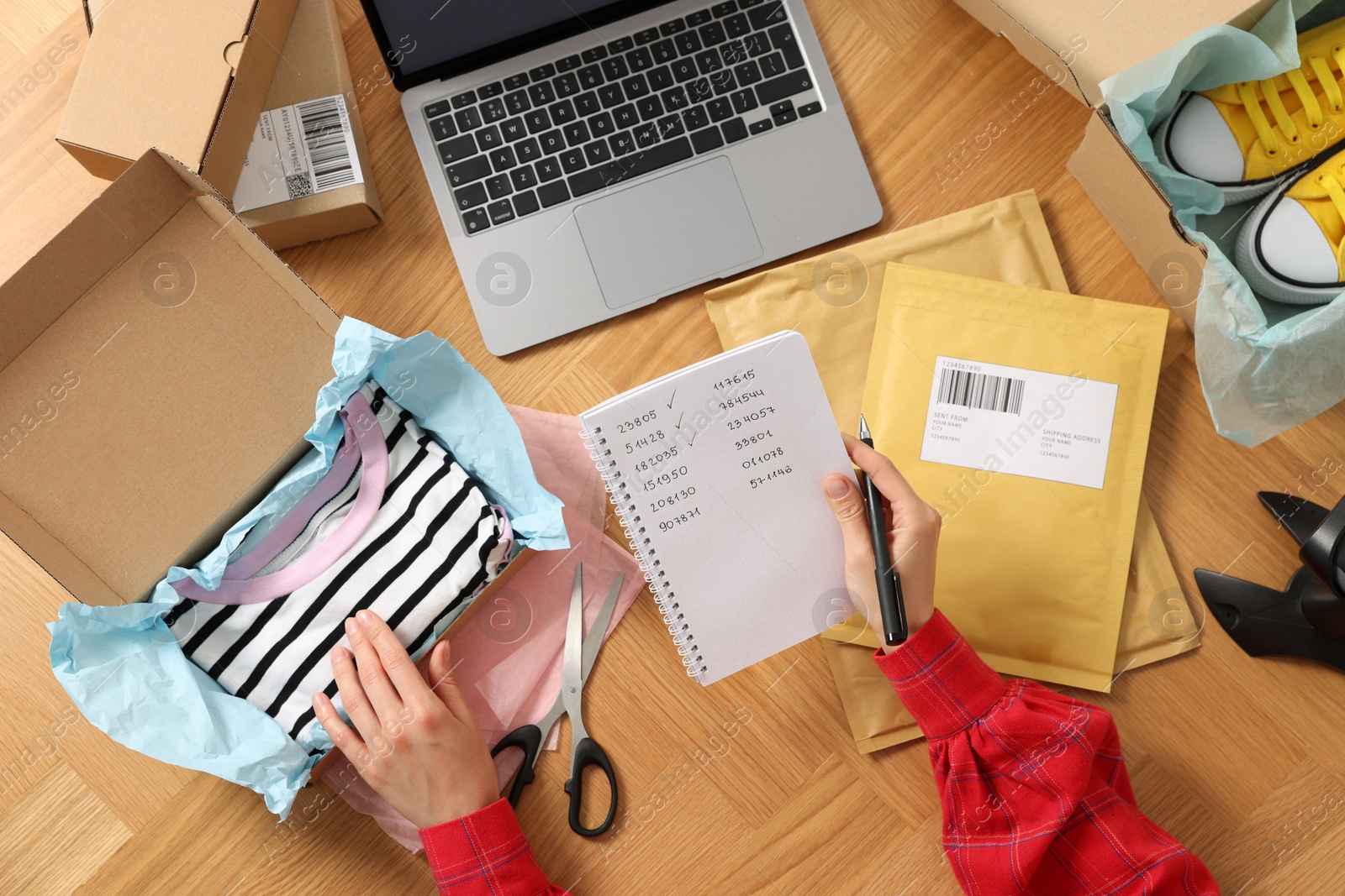 Photo of Seller with parcel and notebook working at wooden table, top view. Online store