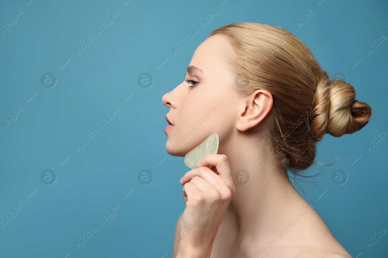 Photo of Beautiful young woman doing facial massage with gua sha tool on blue background