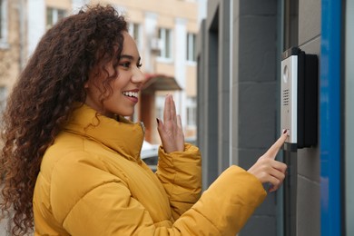 Young African-American woman ringing intercom while waving to camera near building entrance