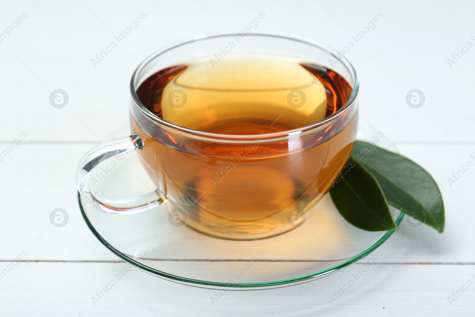 Photo of Aromatic tea in glass cup and green leaves on white wooden table