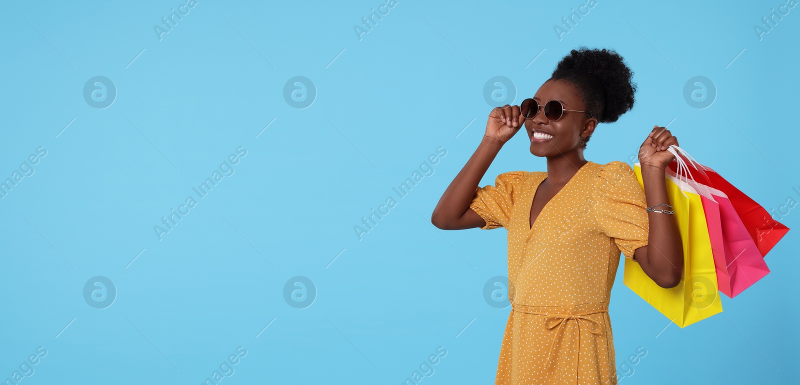 Photo of Happy young woman in stylish sunglasses with shopping bags on light blue background. Space for text