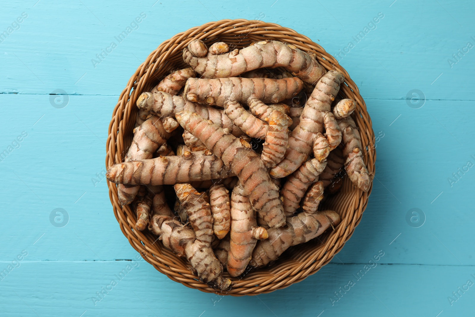 Photo of Wicker bowl with raw turmeric roots on light blue wooden table, top view