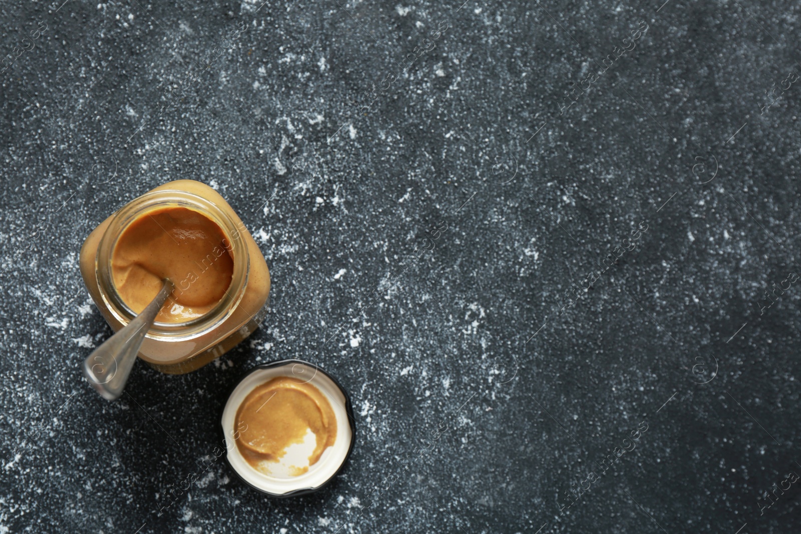 Photo of Spicy mustard and spoon in glass jar on dark grey table, top view. Space for text