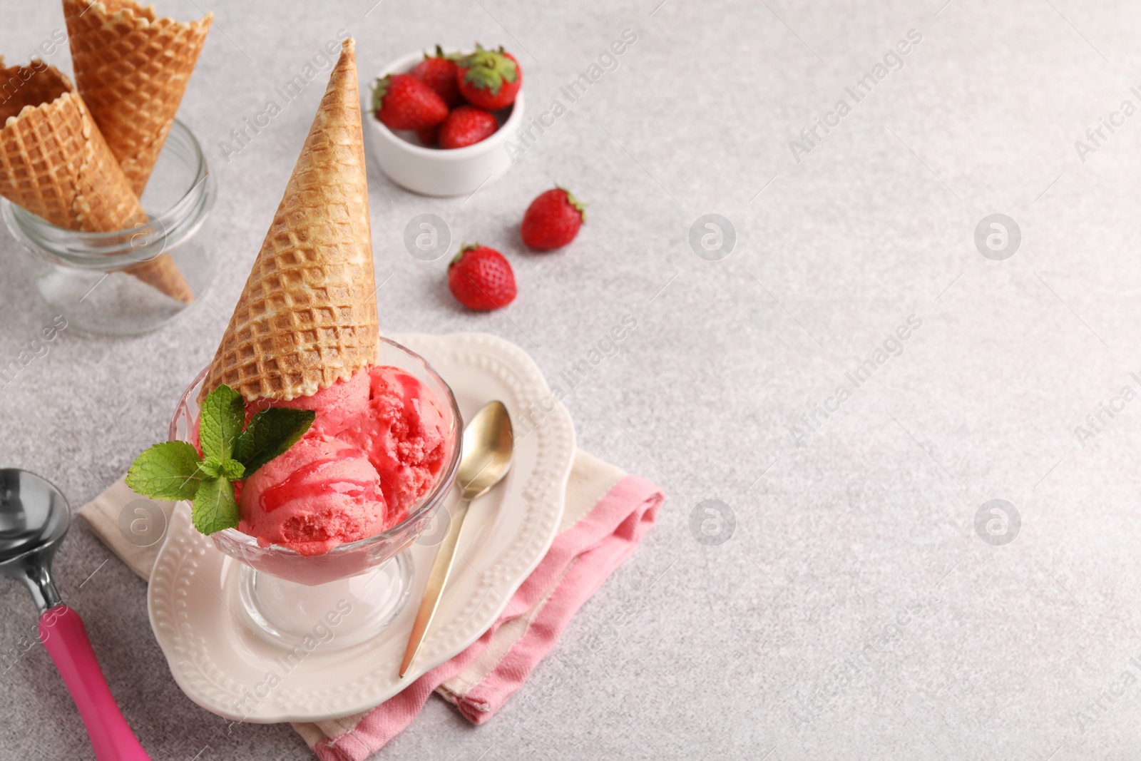 Photo of Delicious scoops of strawberry ice cream with mint and wafer cone in glass dessert bowl served on grey table. Space for text