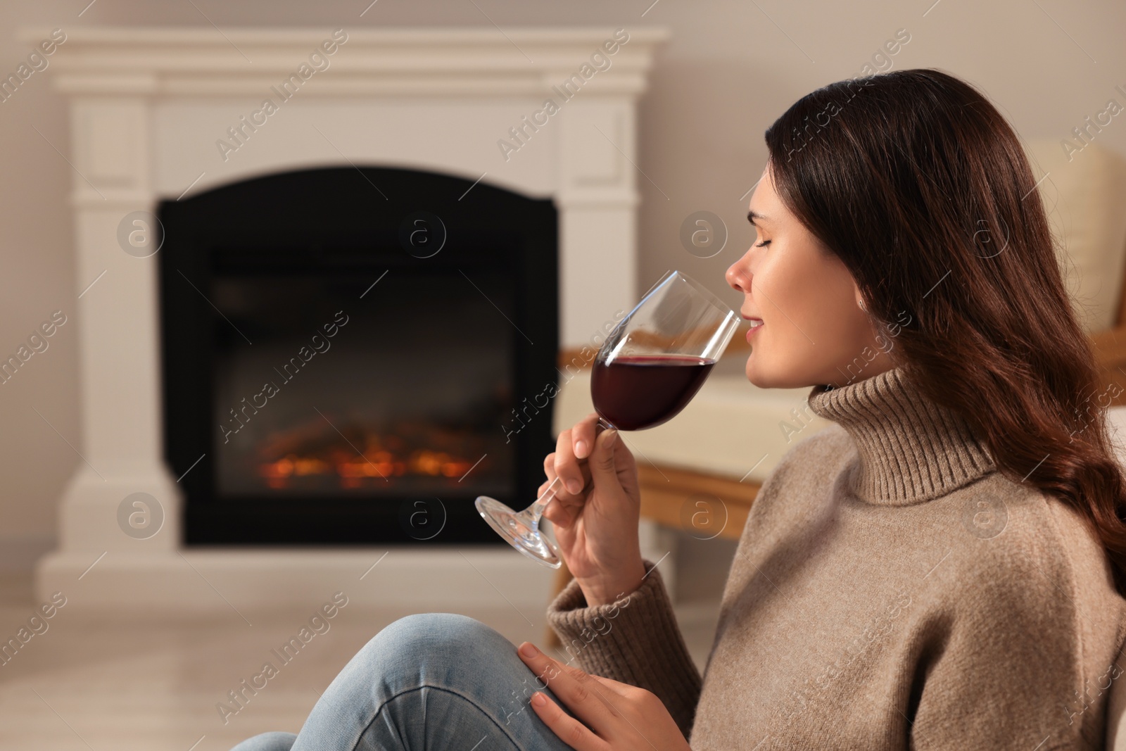 Photo of Young woman with glass of wine relaxing near fireplace at home. Space for text