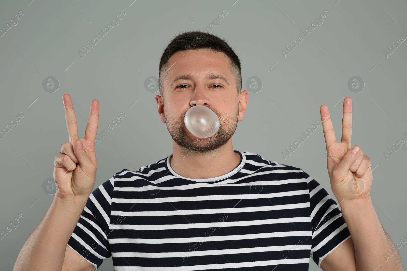 Photo of Handsome man blowing bubble gum and showing peace gesture on light grey background