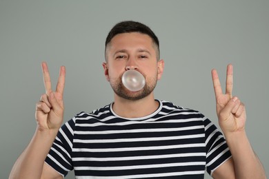 Photo of Handsome man blowing bubble gum and showing peace gesture on light grey background