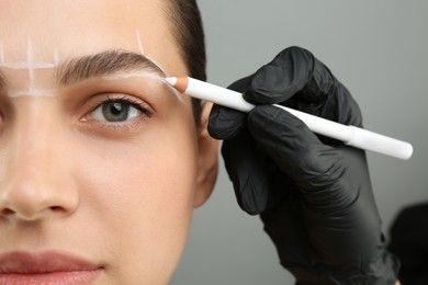 Photo of Beautician preparing young woman for procedure of permanent eyebrow makeup on grey background, closeup