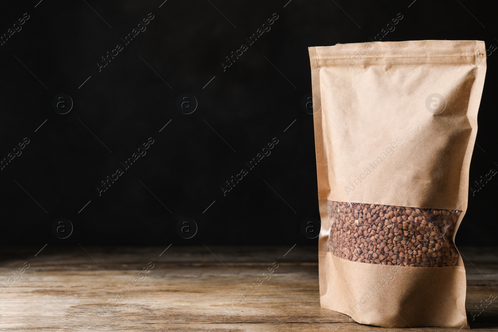 Photo of Buckwheat grains in package on wooden table against black background. Space for text