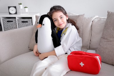 Photo of Little girl dressed as doctor hugging toy penguin on sofa indoors. Pediatrician practice
