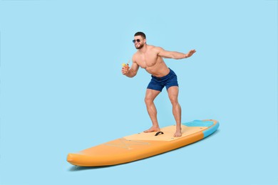 Photo of Happy man with refreshing drink balancing on SUP board against light blue background
