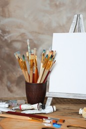 Many brushes, easel with blank canvas and paints on wooden table