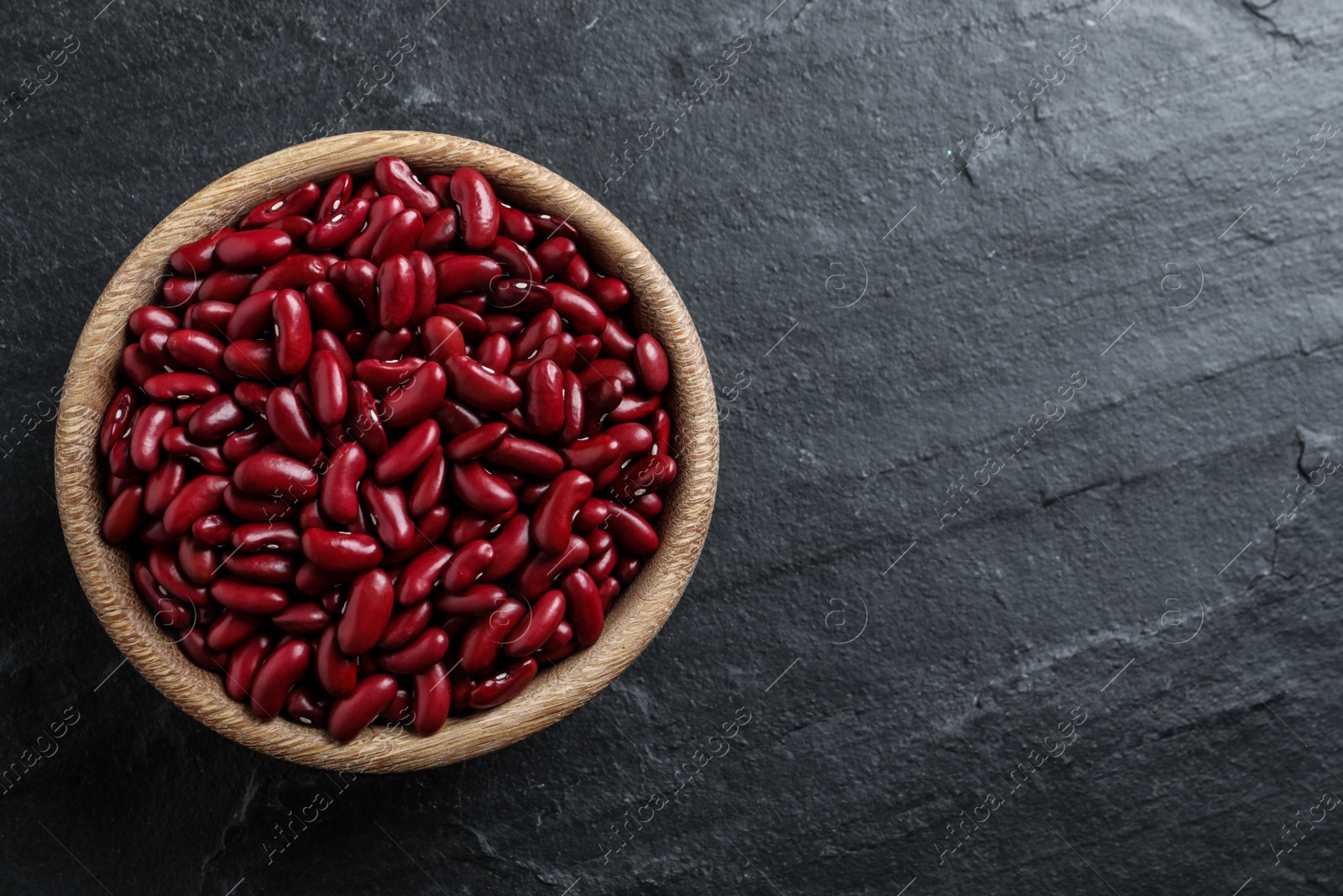 Photo of Raw red kidney beans in wooden bowl on black table, top view. Space for text