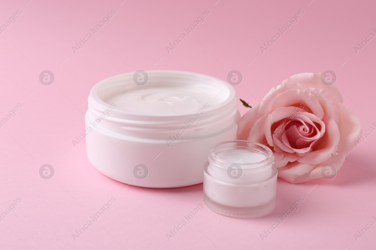 Photo of Moisturizing cream in open jars and rose flower on pink background. Body care product