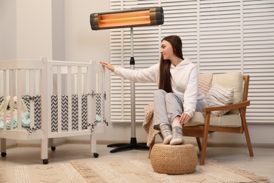 Young woman near crib and modern electric infrared heater indoors