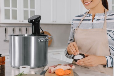 Photo of Woman salting meat near pot with sous vide cooker at table in kitchen, closeup. Thermal immersion circulator