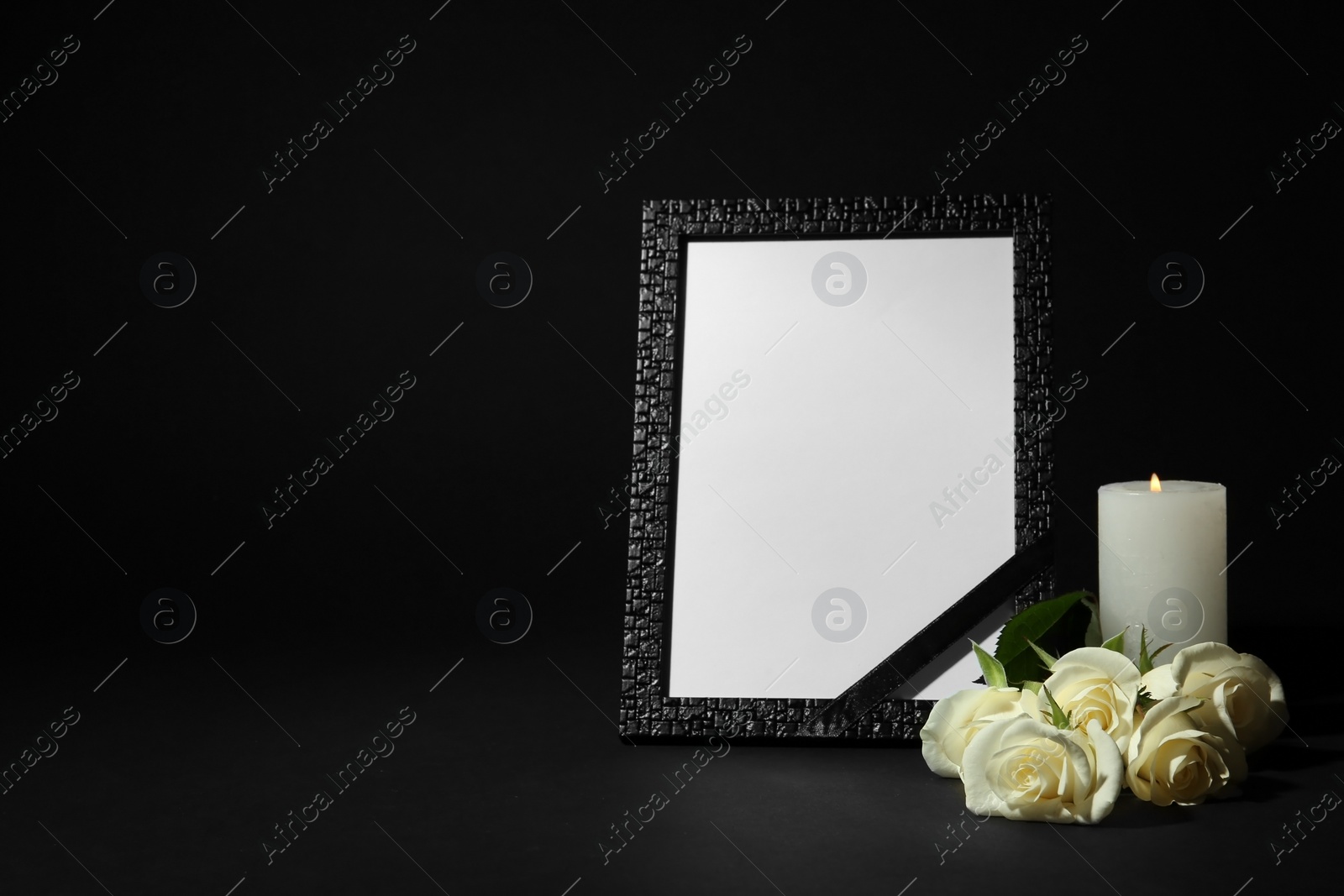 Photo of Funeral photo frame with ribbon, white roses and candle on dark table against black background. Space for design