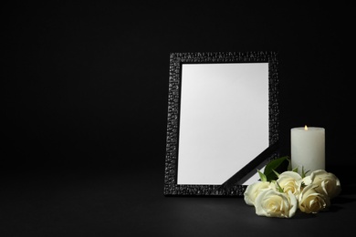 Photo of Funeral photo frame with ribbon, white roses and candle on dark table against black background. Space for design